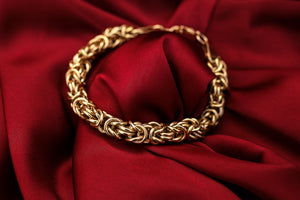 Gold chain bracelet with intricate interwoven links, elegantly displayed on rich red satin fabric.