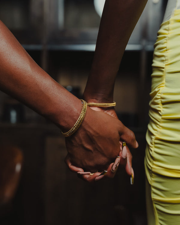 Two hands holding each other with gold bracelets and rings, one wearing a yellow dress.